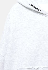 Szürke, rövidített kapucnis pulóver texturált anyagból, melynek kapucniján zsinór van, és fekete „STUDIO” felirat díszíti a nyílást. Fodros hemline.