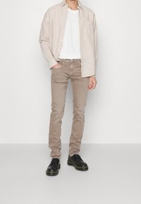 Beige randig skjorta över en vit T-shirt, kombinerad med ljusbruna jeans och svarta skor med gul sömnad. Enkel design och avslappnad passform.