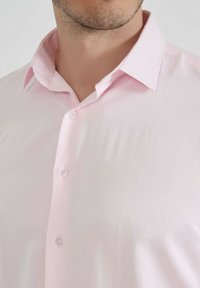 Homme portant une chemise rose clair à boutons, avec un col ouvert, montrant une partie de son cou et une barbe de trois jours.