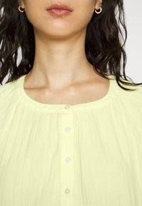 Blusa amarelo claro com decote redondo, textura plissada e cinco pequenos botões na parte da frente. O material parece suave e leve.