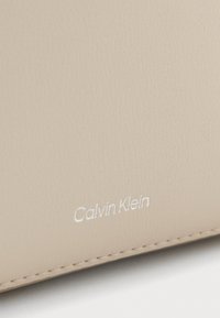 Svetlo bež usnjena denarnica z vidnim šivanjem. Opremljena z embosiranim besedilom "Calvin Klein" v beli barvi. Gladka tekstura in minimalistična zasnova.