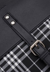 Black synthetic leather strap with silver hardware, fastening over a black and white plaid pattern fabric background. Textured surface visible.