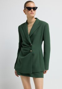 Vihreä blazer, jossa on liehuva muotoilu, yksittäinen kultanappi ja vastaava minihame, kerroksittain beige ribbed neuleen päällä. Mustat kissankäpäläaurinkolasit.