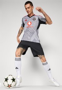 Maglietta da calcio grigia e nera con motivo geometrico, dettagli rossi e logo; abbinata a pantaloni corti neri e calze bianche. Palla da calcio nei dintorni.