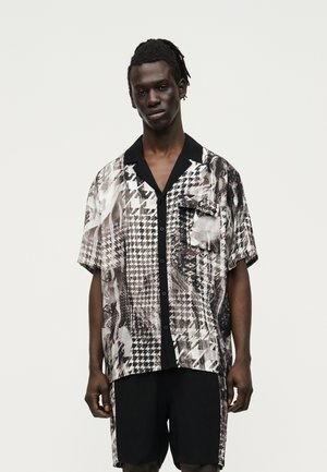 Homme à la peau foncée et aux cheveux tressés, portant une chemise à manches courtes à boutons avec un motif noir et blanc et un short noir avec des panneaux latéraux assortis.