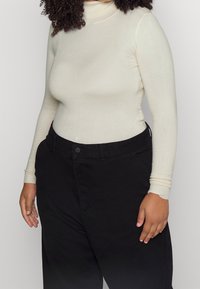 Maglia beige chiaro a maniche lunghe con colletto alto abbinata a pantaloni neri a vita alta. La maglia presenta un design aderente e polsini a coste.