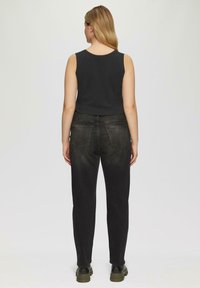 Zwarte mouwloze top gecombineerd met donkere denim jeans, met achterzakken en een taps toelopende pijp. Model staat met de rug naar de camera, met zwarte laarzen aan.