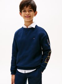 Felpa blu navy con scollatura rotonda e motivi floreali ricamati sulle maniche. Indossata sopra una camicia bianca con colletto, tessuto morbido.