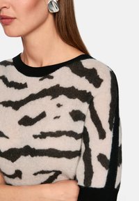 Leichter, durchsichtiger Pullover mit cremefarbenem Grundton und kräftigem schwarzem Tiermuster. Schwarzer, gerippter Halsausschnitt und Kanten ergänzen das Design.