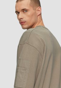 Kaki pullover trui van textuurstof; met een ronde hals en een patchzak op de linkermouw. Losse pasvorm.