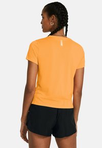 Mujer con el cabello trenzado que lleva una camisa de manga corta naranja y pantalones cortos de deporte negros, mostrada de espaldas contra un fondo liso.