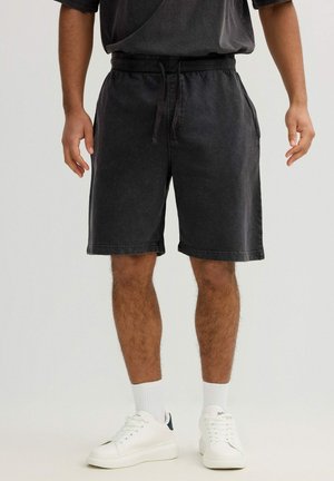 Hombre con pantalones cortos negros con cordón, calcetines blancos y zapatillas blancas de pie contra un fondo liso.