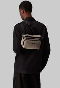 Beige szövetből készült keresztpántos táska cipzáros elülső zsebbel, állítható pánttal és struktúrált dizájnnal, fekete felsőruházat fölött viselve.