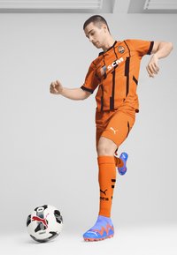Equipación de fútbol naranja con rayas negras y logo; botas de fútbol azules con detalles en rosa; balón de fútbol en blanco y negro con logo y patrones.