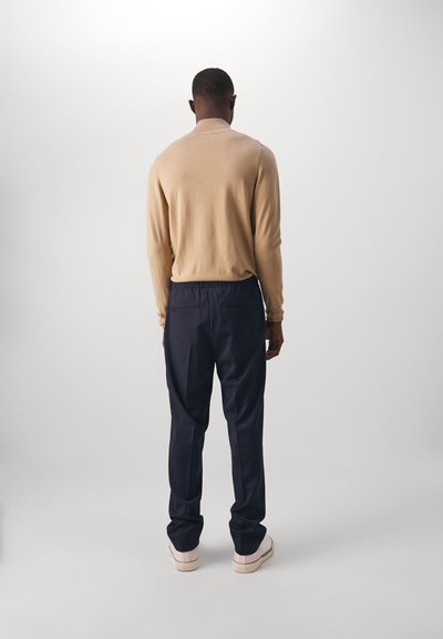 Beige langærmet sweater med høj krave, parret med marineblå bukser med elastisk talje og lyse sko. Set fra ryggen.