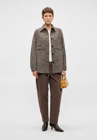 Sieviete ar īsiem matiem, ģērbusies brūnā rūtainā žaketē, brūnās biksēs un melnās augstpapēžu kurpēs, tur nelielu sinepju krāsas rokassomiņu.