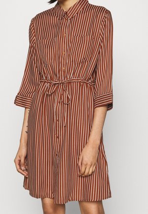 Femme portant une robe marron à rayures blanches verticales, boutonnée devant, poches poitrine, manches trois-quarts et ceinture nouée à la taille.