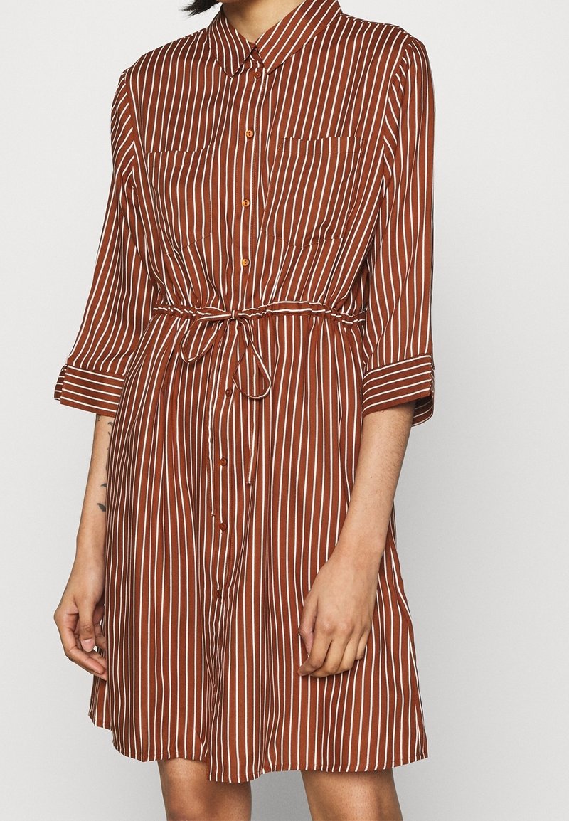 Femme portant une robe marron à rayures blanches verticales, boutonnée devant, poches poitrine, manches trois-quarts et ceinture nouée à la taille.