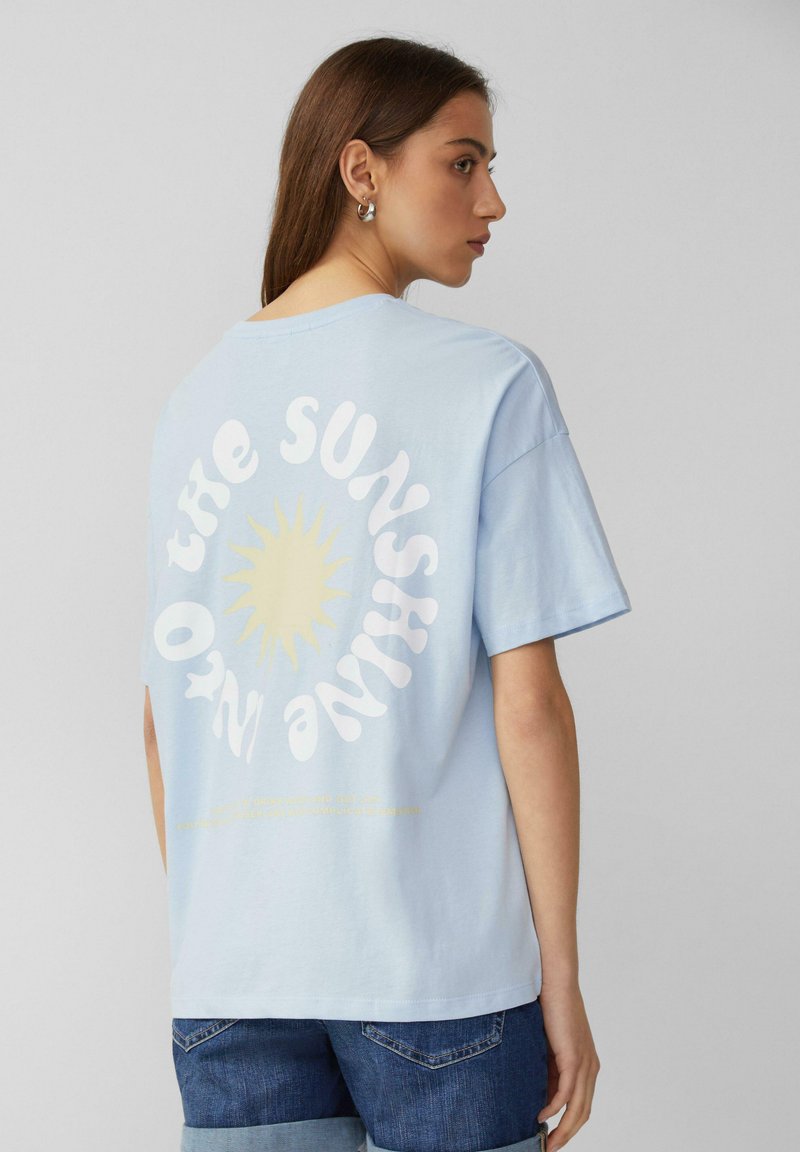 Lichtblauwe katoenen t-shirt met een zonafbeelding en de tekst "DE ZON" in het wit. Korte mouwen en een ontspannen pasvorm. Gecombineerd met denim shorts.