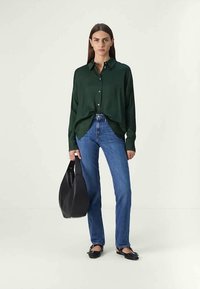 Mulher vestindo uma camisa de botão verde-escuro, jeans azuis e sapatos pretos, segurando uma grande bolsa preta, pousando contra um fundo simples.
