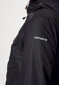 Schwarze isolierte Jacke mit elegantem Design, ausgestattet mit hohem Kragen, verstellbarer Kapuze und einem weißen "ICEPEAK"-Logo am Ärmel.
