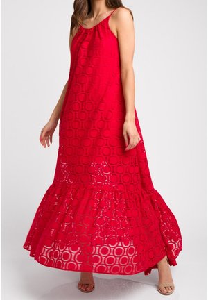 Kvinde iført rød maxikjole uden ærmer med cirkulært blondermønster og flæsekant, kombineret med beige sandaler med åbne tæer.