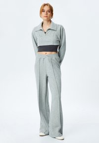 Grijze cropped sweatshirt met een brede kraag, ritssluiting en grijze high-waisted broek, beide in een zachte, gestructureerde stof. Witte schoenen zijn zichtbaar.