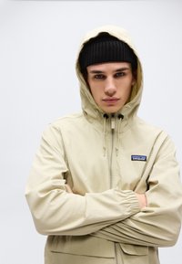 Νεαρός άνδρας με μπεζ μπουφάν με κουκούλα και μαύρο σκουφάκι, χέρια σταυρωμένα, κοιτάζει κατευθείαν μπροστά σε απλό ανοιχτό φόντο.