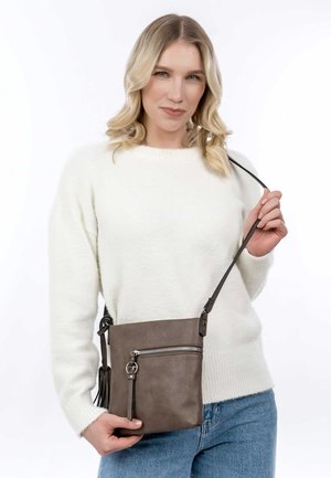Sac en bandoulière en cuir marron avec fermeture éclair argentée, un détail de frange sur le côté et une sangle réglable, porté par une personne vêtue d'un pull blanc.
