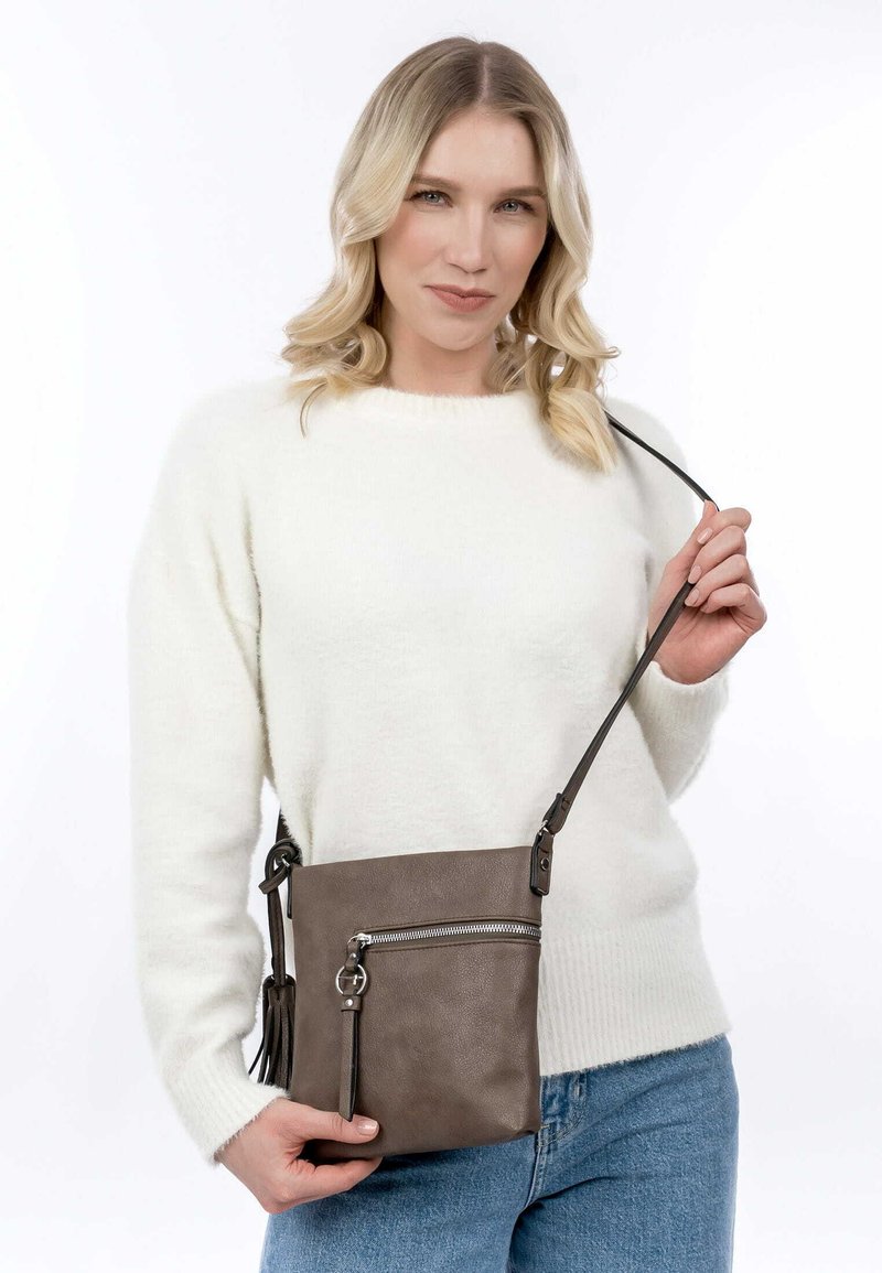 Hnědá kožená crossbody taška se stříbrným zipem, postranním třásňovým detailem a nastavitelným popruhem, kterou drží osoba v bílém svetru.