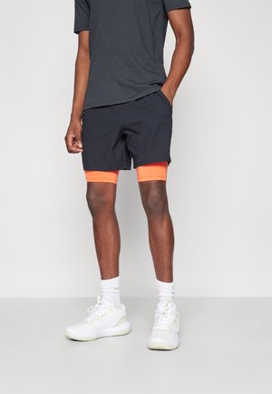 Mężczyzna ubrany w czarne spodenki na jasnopomarańczowych spodenkach kompresyjnych, szary t-shirt, białe skarpetki i białe buty sportowe, stojący z rękami w kieszeniach.