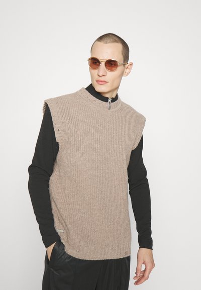 Άνδρας φορεμένος με μπεζ αμάνικο πλεκτό πουλόβερ πάνω από μαύρο πουκάμισο με μακριά μανίκια, μαύρο παντελόνι και στρογγυλά καφέ γυαλιά ηλίου, ποζάροντας με τα χέρια του partially στις τσέπες.