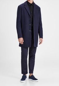 Marineblauwe jas van een wollen mix met inkeping revers, drie knopen en zijzakken, gecombineerd met donkere broek en marineblauwe sneakers, allemaal in een minimalistische stijl.