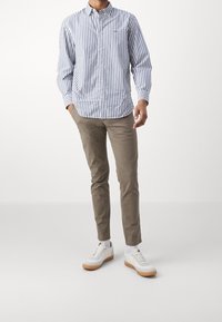 Pruhovaná košile na knoflíky v modro-bílé barvě, spárovaná se béžovými slim-fit kalhotami a bílými teniskami s hnědými podrážkami. Čistý, funkční design.