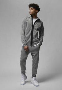 Tuta sportiva grigia con giacca con cappuccio e zip e pantaloni affusolati. Presenta il logo Jordan, strisce laterali e sneaker bianche.