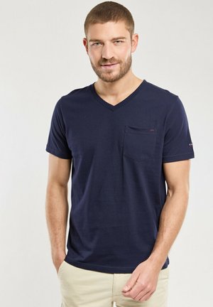 T-shirt bleu marine en V en tissu doux, avec une poche poitrine et un logo discret sur la manche. Texture lisse, coupe décontractée.