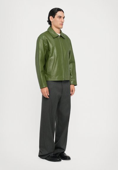 Chaqueta de cuero verde con cuello de punta y cierre de cremallera, combinada con pantalones de pierna ancha grises y zapatos negros. Diseño sencillo, textura suave.