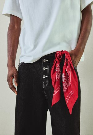 Osoba nosiaca čierne rifle s kontrastným prešívaním a štyrmi gombíkmi, biele tričko a červenú vzorovanú bandanu visiacu z vrecka.