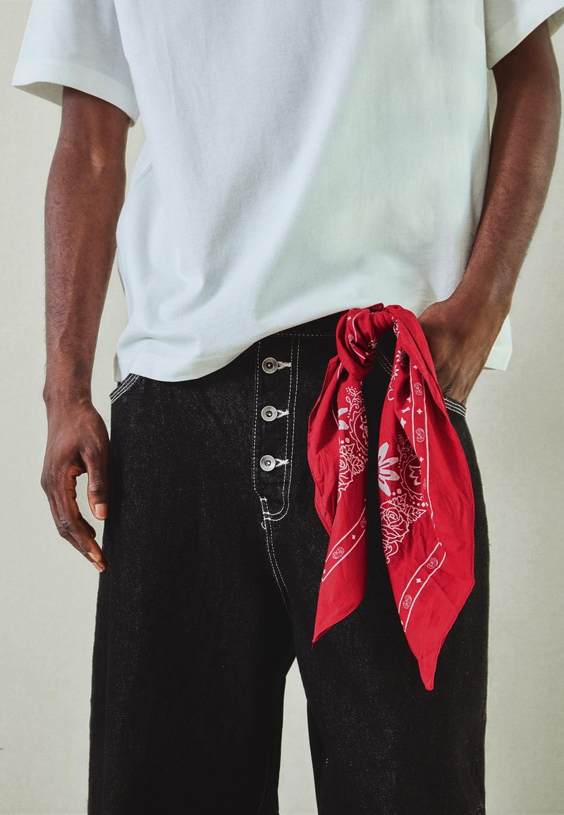 Personne portant un jean noir avec des coutures contrastantes et quatre boutons, un t-shirt blanc, et un bandana rouge à motif suspendu à la poche.