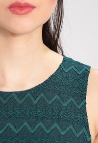 Robe en dentelle teal avec un col rond. Présente des motifs en zigzag et floraux, un tissu texturé et une silhouette ajustée.