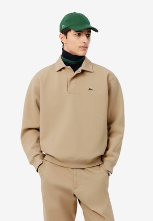 Giovane uomo che indossa una felpa beige e pantaloni con berretto verde, in piedi con le mani in tasca, leggermente rivolto a destra su sfondo bianco.