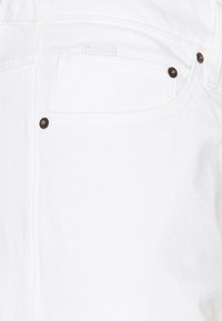 Tejido de mezclilla blanca, con un bolsillo trasero que presenta una pequeña etiqueta, remaches de cobre y una textura limpia y suave. Sin patrones ni acentos visibles.