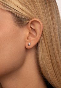 Pendiente de plata con un diseño triangular, que presenta un centro negro y un pequeño acento de piedra clara. Acabado suave, tamaño compacto.