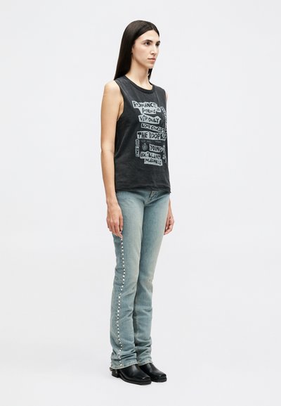 Woman with long dark hair wearing a sleeveless black graphic top, light blue jeans with side studs, and black ankle boots, standing against white background.