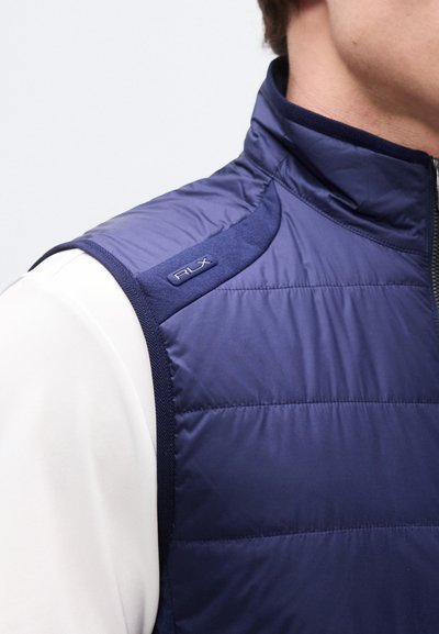 Άνδρας που φοράει μπλε σκούφο αμάνικο μπουφάν με καπιτονέ σχέδιο πάνω από λευκό πουκάμισο με μακριά μανίκια, εμφανίζοντας το πάνω μέρος του κορμού και λεπτομέρειες για το γιακά με το λογότυπο "RLX".