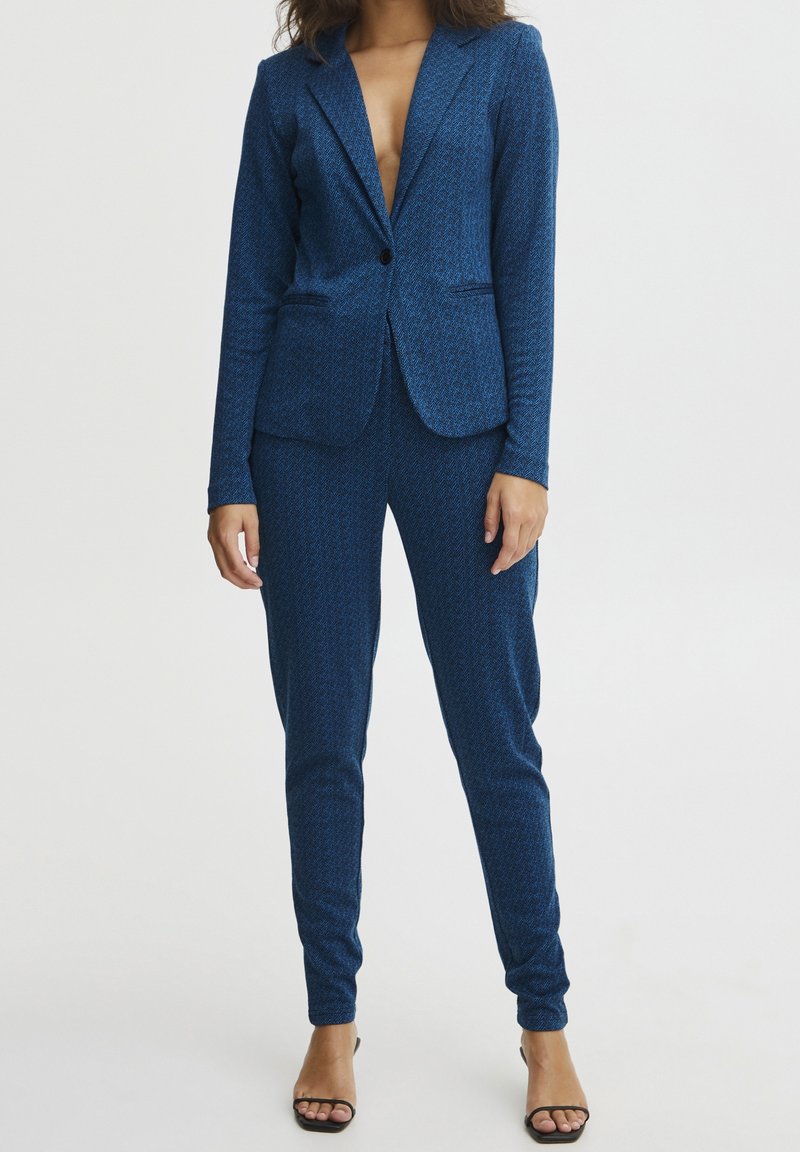 Modrý vzorovaný oblek s přiléhavým střihem, obsahující upravený blejzr a zúžené kalhoty, vyrobený z texturované látky. Má jednořadé zapínání na knoflík a klopové límce.