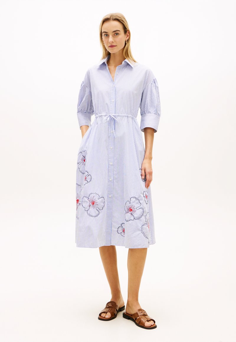 Femme debout portant une robe chemise midi rayée bleu et blanc avec broderie florale, manches trois-quarts, et sandales marron à bout ouvert.