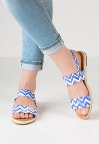 Blå og hvide chevron-mønstrede sandaler med to brede remme, flad sål og teksturerede kanter. Båret med lyseblå ankel-lange jeans.