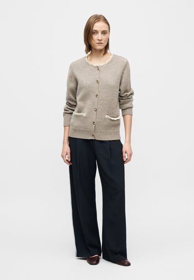 Casaco de malha castanho claro com textura canelada, apresentando um decote com bordas onduladas, botões na frente e bolsos de remendo com detalhes em creme.