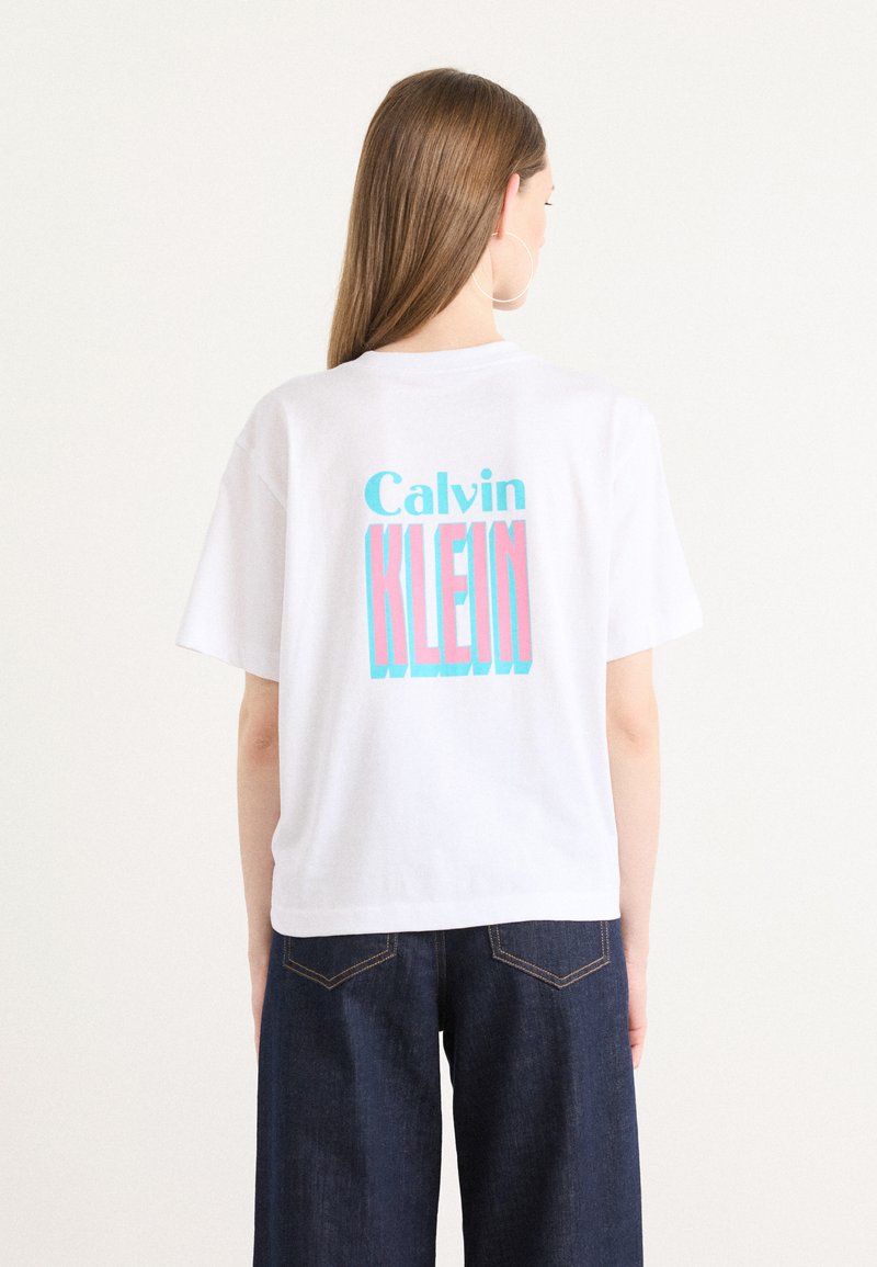 T-shirt en coton blanc avec un graphique coloré "Calvin KLEIN" en rose et bleu au dos. Coupe décontractée avec des manches courtes.